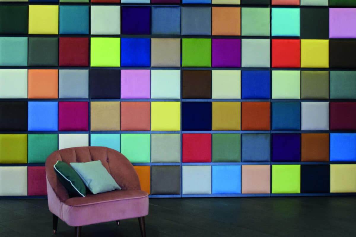Polsterstoff Denver XL – rosa Sessel mit grünen und hellblauen Kissen vor Wand aus bunten Stoffmustern